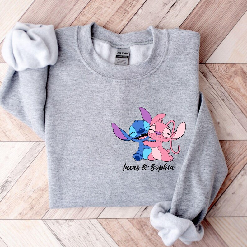 May include: A grey sweatshirt with a cartoon illustration of two characters, one blue and one pink, hugging. The text "Lucas & Sophia" is printed below. The sweatshirt has a crew neck and long sleeves.