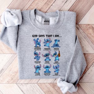 Op de afbeelding: Een lichtgrijs sweatshirt met een afbeelding van het personage Stitch. De tekst "GOD SAYS THAT I AM..." staat boven het personage, met woorden als "EMPOWERED", "TREASURED" en "PRECIOUS" onder elke afbeelding van Stitch.