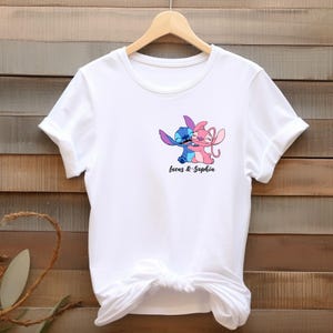 May include: White t-shirt featuring a graphic of two cartoon characters, one blue and one pink, embracing. The text "Lucas & Sophia" is printed below the characters in black script. The shirt has short sleeves and a crew neck.