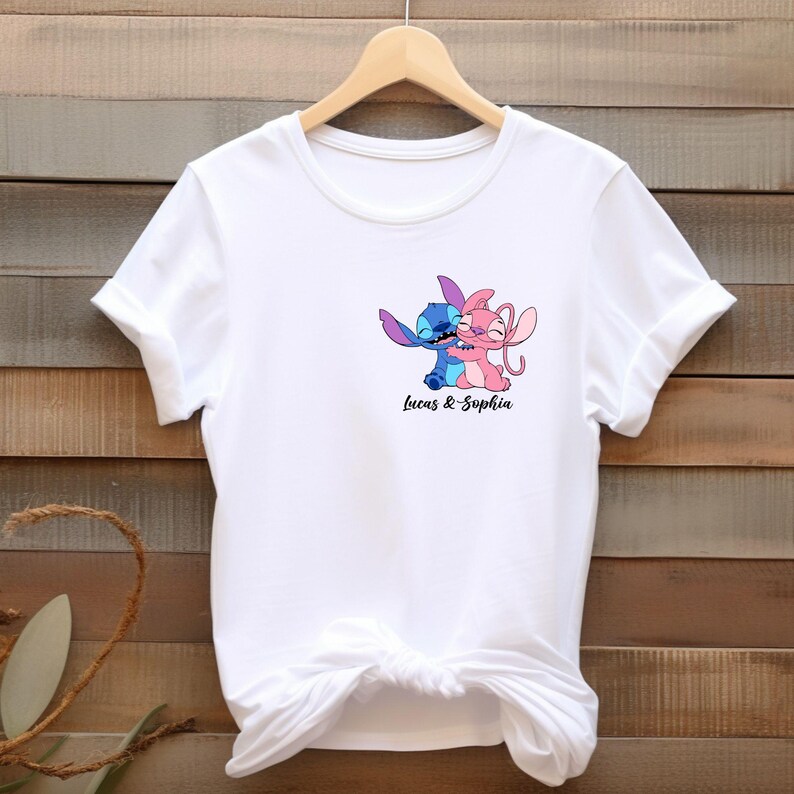May include: White t-shirt featuring a graphic of two cartoon characters, one blue and one pink, embracing. The text "Lucas & Sophia" is printed below the characters in black script. The shirt has short sleeves and a crew neck.