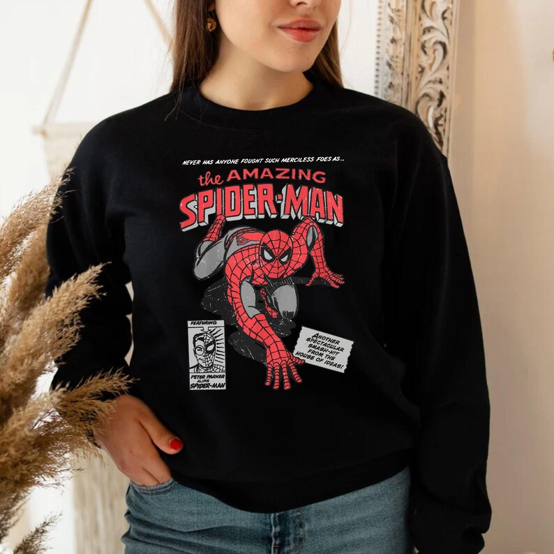 May include: Black sweatshirt featuring a comic book style graphic of Spider-Man in red and gray. The text "the AMAZING SPIDER-MAN" is in red and white. Additional text includes "NEVER HAS ANYONE FOUGHT SUCH MERCILESS FOES AS..." and other comic book elements.