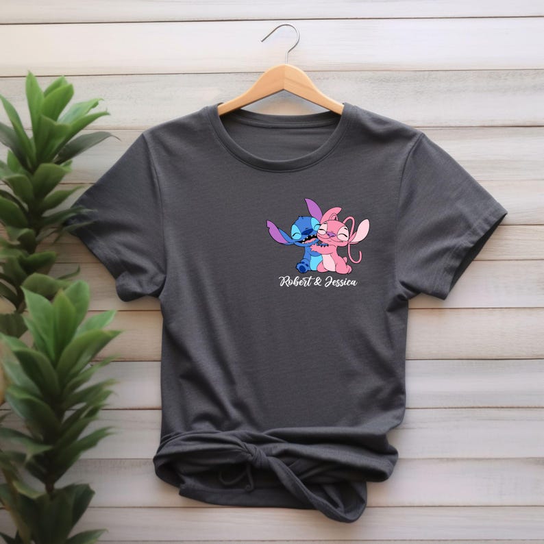 May include: Dark gray t-shirt featuring a cartoon illustration of two characters embracing, one blue and one pink. The text "Robert & Jessica" is printed below the graphic. The shirt is hanging on a wooden hanger.