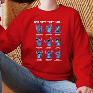 Op de afbeelding: Een rode sweatshirt met een afbeelding van het personage Stitch en de tekst "GOD SAYS THAT I AM..." samen met positieve beweringen zoals "Empowered", "Treasured" en "Precious". Het ontwerp bevat bijbelverzen.