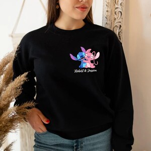 May include: Black crew neck sweatshirt with a colourful graphic of two cartoon characters hugging. The characters are blue and pink, with the text "Robert & Jessica" beneath. The sweatshirt is a casual style.