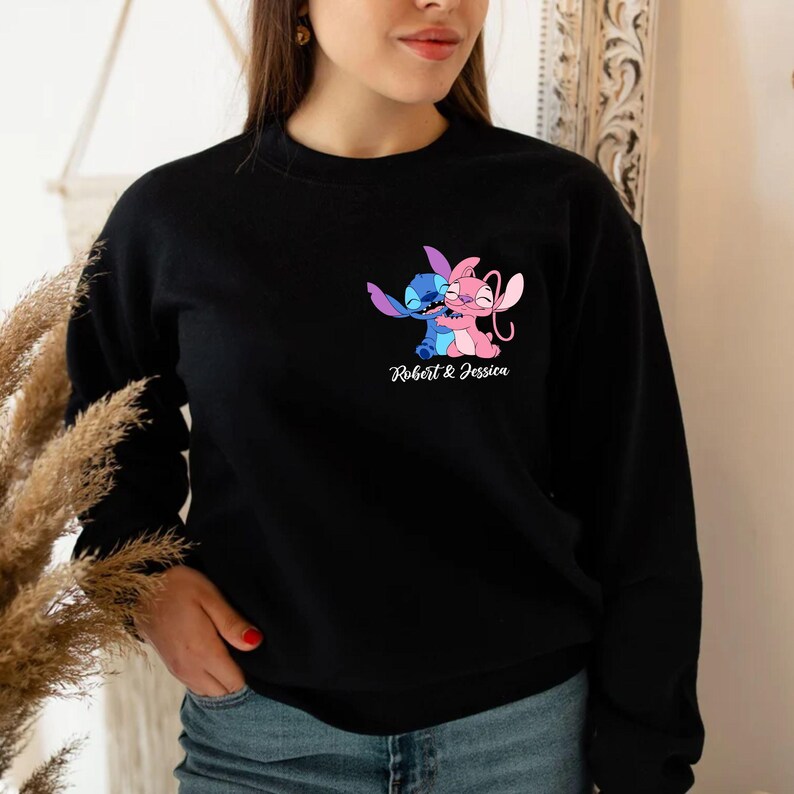 May include: Black crew neck sweatshirt with a colourful graphic of two cartoon characters hugging. The characters are blue and pink, with the text "Robert & Jessica" beneath. The sweatshirt is a casual style.