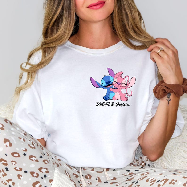 May include: White t-shirt featuring a cartoon illustration of two characters embracing, one blue and one pink. The text "Robert & Jessica" is below the illustration. The shirt is worn by a person.