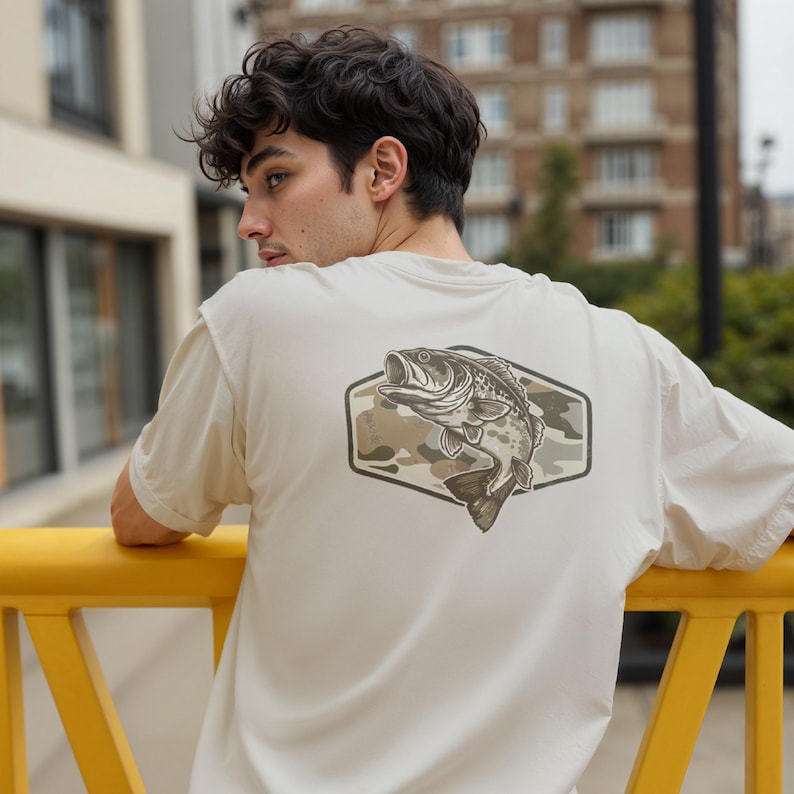 May include: A cream-coloured t-shirt with a graphic of a fish leaping from water, set against a camouflage background within a hexagonal frame. The shirt has short sleeves and a neutral tone.