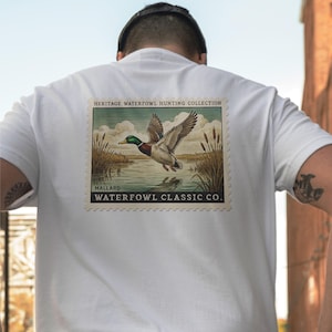 May include: White t-shirt featuring a vintage-style graphic of a mallard duck in flight over water, with cattails and a landscape background. The graphic includes the text "HERITAGE WATERFOWL HUNTING COLLECTION" and "WATERFOWL CLASSIC CO."