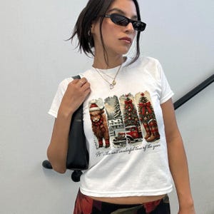 May include: White t-shirt with a festive graphic featuring a Highland cow in a Santa hat, Christmas trees, a red truck, and plaid boots. The text "It's the most wonderful time of the year" is printed below the images.
