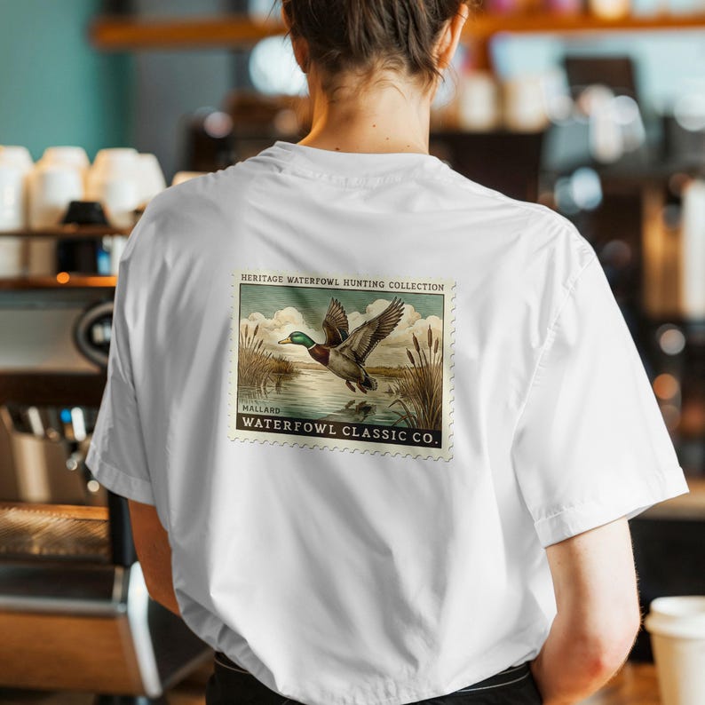 May include: White short-sleeve shirt with a graphic on the back. The graphic is a vintage-style illustration of a mallard duck in flight over water, with the text "HERITAGE WATERFOWL HUNTING COLLECTION" and "WATERFOWL CLASSIC CO."