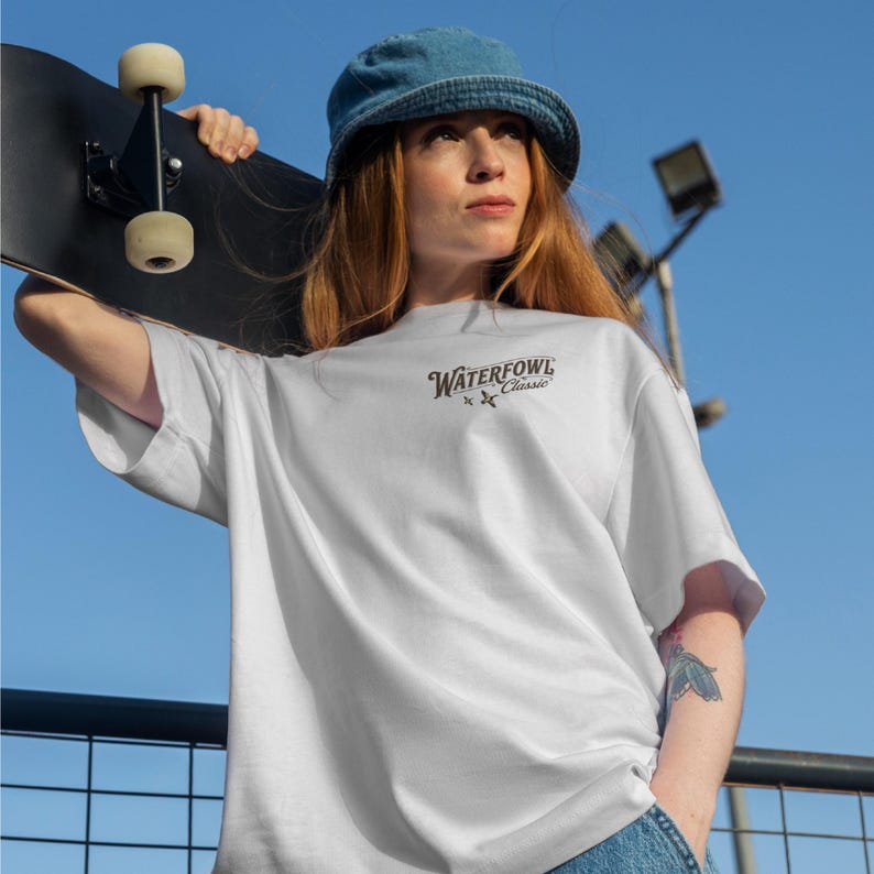 May include: White oversized t-shirt with the text "Waterfowl Classic" in a dark font. The shirt is worn with a denim bucket hat and blue jeans. A black skateboard is held in the background. The sky is blue.