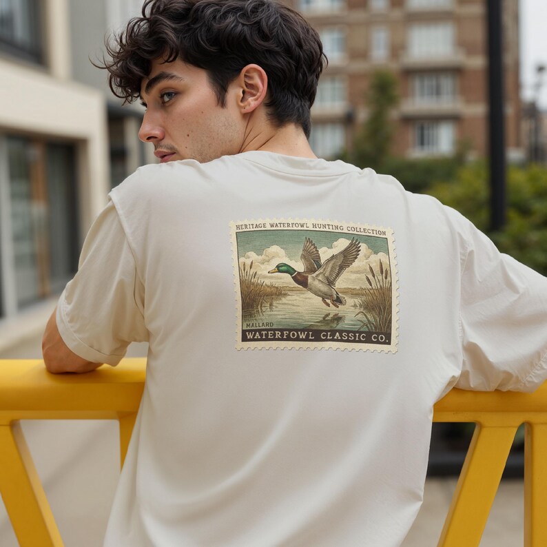 May include: A light beige t-shirt featuring a vintage-style graphic of a mallard duck in flight over a lake. The graphic is framed with text that reads "HERITAGE WATERFOWL HUNTING COLLECTION" and "WATERFOWL CLASSIC CO."