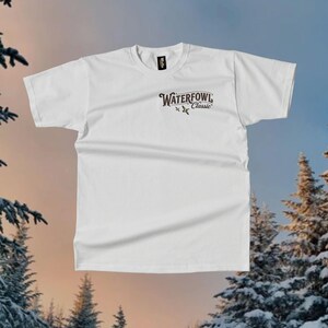 May include: A white t-shirt featuring the text "Waterfowl Classic" in a brown, stylized font. The shirt has a crew neck and short sleeves. The background shows a winter scene with snow-covered trees.