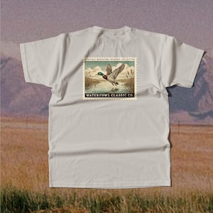 May include: A light gray t-shirt featuring a vintage-style graphic of a mallard duck in flight over water. The graphic is framed by a postage stamp design with the text "HERITAGE WATERFOWL HUNTING COLLECTION" and "WATERFOWL CLASSIC CO."
