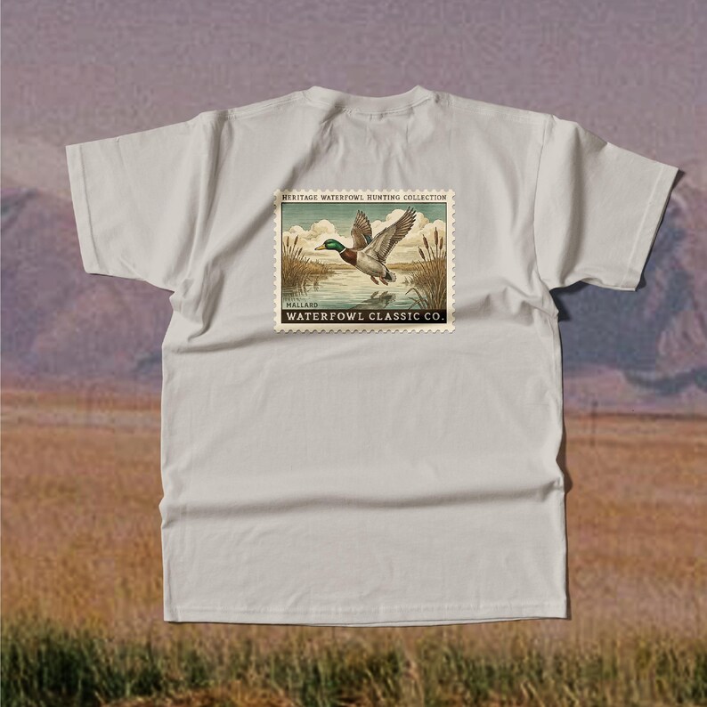 May include: A light gray t-shirt featuring a vintage-style graphic of a mallard duck in flight over water. The graphic is framed by a postage stamp design with the text "HERITAGE WATERFOWL HUNTING COLLECTION" and "WATERFOWL CLASSIC CO."