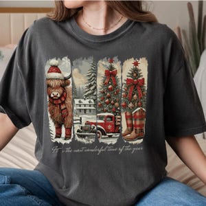 May include: Dark gray t-shirt featuring a winter scene with a Highland cow in a hat and scarf, a snowy house, a red truck, and Christmas trees. The text "It's the most wonderful time of the year" is printed at the bottom.