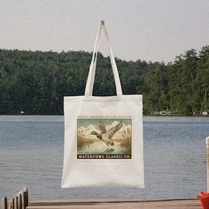 May include: A white tote bag with a vintage-style illustration of a mallard duck in flight over water. The bag features the text "HERITAGE WATERFOWL HUNTING COLLECTION" and "WATERFOWL CLASSIC CO."