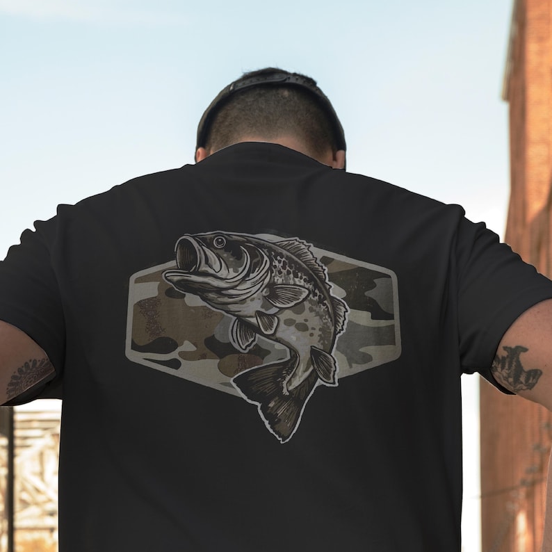 May include: Black t-shirt with a detailed illustration of a largemouth bass. The fish is set against a camouflage background within a hexagonal shape. The design is centered on the back of the shirt.