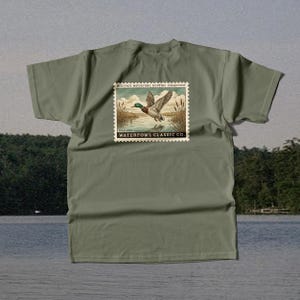 May include: An olive green t-shirt featuring a vintage-style stamp design. The stamp depicts a mallard duck in flight over water, with the text "HERITAGE WATERFOWL HUNTING COLLECTION" and "WATERFOWL CLASSIC CO."