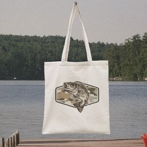 May include: Cream-coloured tote bag with a fish illustration inside a camouflage hexagon. The bag has long handles and is set against a lake and trees. The fish is in shades of grey and brown. Ideal for carrying items.
