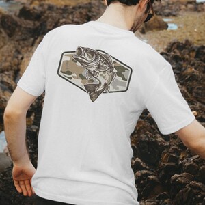 May include: White t-shirt with a fish illustration on the back. The fish is within a camouflage-patterned, geometric shape. The fish is in shades of grey and brown, with its mouth open. The front of the shirt is plain.
