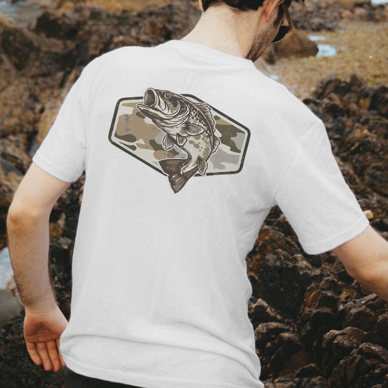 May include: White t-shirt with a fish illustration on the back. The fish is within a camouflage-patterned, geometric shape. The fish is in shades of grey and brown, with its mouth open. The front of the shirt is plain.