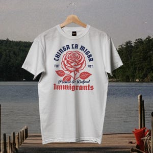 May include: White t-shirt with a red rose graphic and the text "Chinga La Migra" in blue, with "Protect & Defend Immigrants" in red. The shirt is hanging on a wooden hanger.