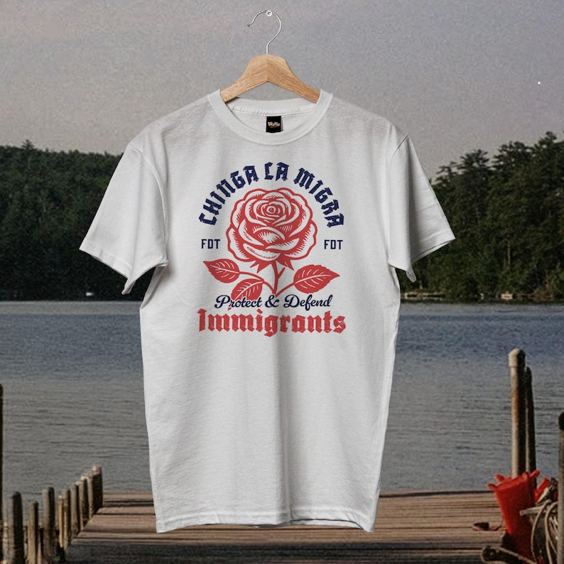 May include: White t-shirt with a red rose graphic and the text "Chinga La Migra" in blue, with "Protect & Defend Immigrants" in red. The shirt is hanging on a wooden hanger.