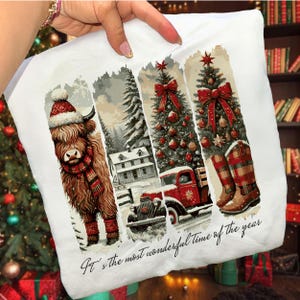 May include: White pillow with a winter scene design. The design includes a Highland cow in a Santa hat and scarf, a snowy landscape, a red truck, Christmas trees, and plaid boots. The text reads "It's the most wonderful time of the year."