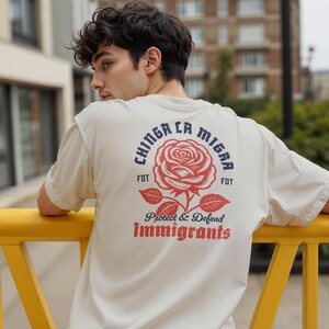 May include: Off-white t-shirt with a red rose graphic and text that reads "CHINGA LA MIGRA" above the rose and "Protect & Defend Immigrants" below. The shirt has short sleeves and a relaxed fit.