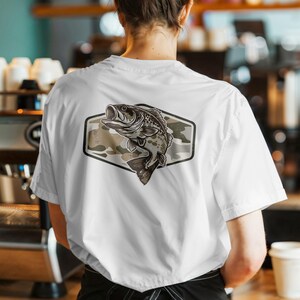 May include: White t-shirt with a fish illustration inside a camouflage-patterned, hexagonal design. The fish is in grey and brown, the camouflage in muted green and tan. The shirt has short sleeves. The design is on the back of the shirt.