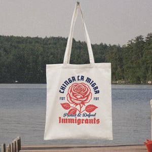 May include: Cream-colored tote bag with a red rose graphic and the text "CHINGA LA MIGRA" in blue, and "Protect & Defend Immigrants" in red. The bag has long white handles and is hanging against a lake backdrop.