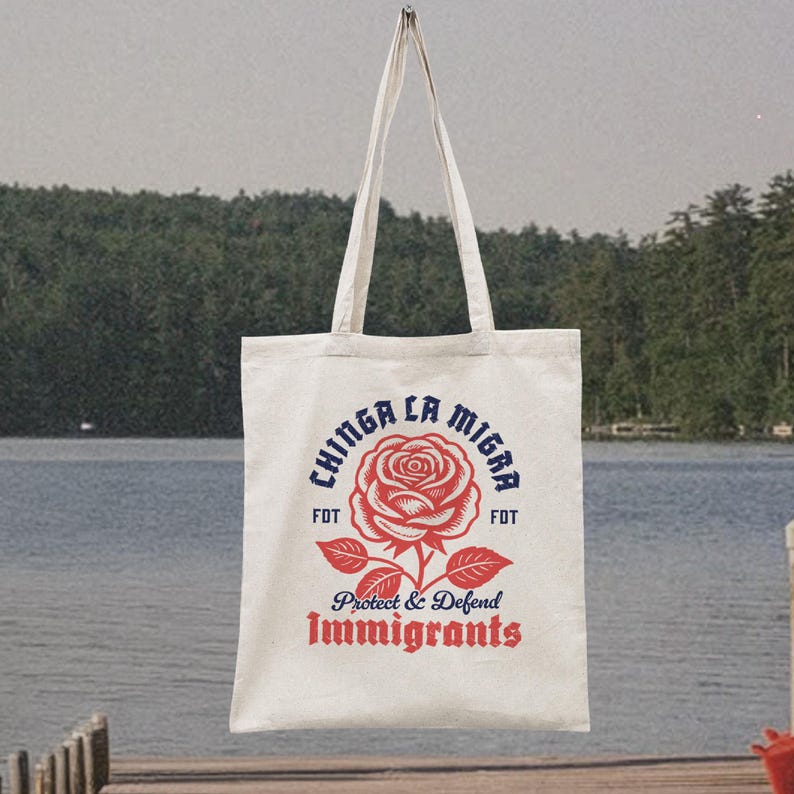 May include: Cream-colored tote bag with a red rose graphic and the text "CHINGA LA MIGRA" in blue, and "Protect & Defend Immigrants" in red. The bag has long white handles and is hanging against a lake backdrop.
