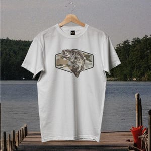 May include: White t-shirt with a graphic of a largemouth bass jumping upwards. The fish is set against a camouflage pattern within a hexagonal frame. The t-shirt is displayed on a wooden hanger.