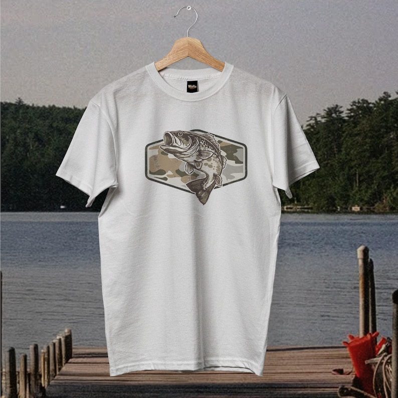 May include: White t-shirt with a graphic of a largemouth bass jumping upwards. The fish is set against a camouflage pattern within a hexagonal frame. The t-shirt is displayed on a wooden hanger.