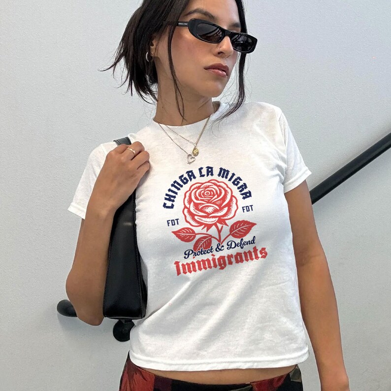 May include: White t-shirt with a graphic design featuring a red rose and the words "Chinga La Migra" and "Protect & Defend Immigrants" in blue and red. The shirt has short sleeves.