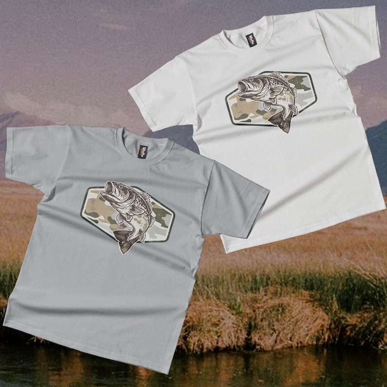 May include: Two short-sleeved t-shirts, one light grey and one white, with a graphic of a largemouth bass leaping from water. The fish is set against a camouflage background within a hexagonal shape. The shirts are casual wear.