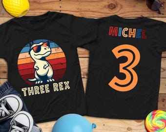 May include: Black t-shirts with a dinosaur graphic. One shirt features a cartoon T-Rex wearing sunglasses with the text "THREE REX". The other shirt has the name "MICHEL" and the number "3" in orange.
