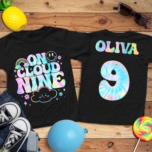 May include: Two black t-shirts with colorful designs. One shirt says "ON CLOUD NINE" with a rainbow, smiley face, and cloud graphic. The other shirt says "OLIVA" and has a tie-dye number "9".