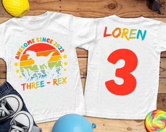 May include: White t-shirts with dinosaur graphics. One shirt features a colorful sunset design with a dinosaur silhouette and the text "AWESOME SINCE 2022 THREE-REX". The other shirt has the name "LOREN" and a large red number "3".