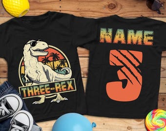 May include: Black t-shirts with dinosaur graphics. One shirt features a dinosaur wearing sunglasses with the text "THREE-REX". The other shirt has the text "NAME" and the number "3". Accessories include sunglasses, a blue balloon, and a lollipop.