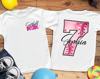 May include: Two white t-shirts. One has "Softball MOM" in pink script. The other has a pink softball-themed number 7 with the name "Sophia" in script. The shirts are on a wooden surface with accessories.