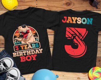 May include: Two black t-shirts. One features a dinosaur wearing sunglasses with the text "3 YEARS BIRTHDAY BOY". The other shirt has the name "JAYSON" and a large number "3" with claw marks. Accessories include sunglasses, a balloon, and a lollipop.