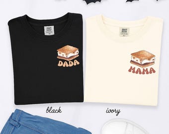 Op de afbeelding: Zwarte en ivoorkleurige crewneck sweatshirts met een s'mores-afbeelding en de woorden "DADA" en "MAMA" in bruin. De sweatshirts worden getoond met een blauwe jeans en witte sneakers.