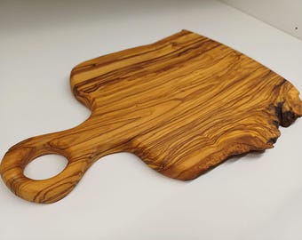 Op de afbeelding: Houten snijplank van olijfhout met een handvat en een gat. Het hout heeft een natuurlijk nerfpatroon in bruine en beige tinten. Het handvat is afgerond en de rand van de plank heeft een unieke, organische vorm. Ideaal voor de keuken.