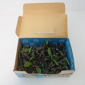 Op de afbeelding: Een open kartonnen doos gevuld met talrijke kleine, donkergroene en zwarte speelgoedsoldaten. De doos heeft een blauwe binnenkant met witte illustraties en een bruine buitenkant. Groen plastic gebladerte is ook aanwezig.