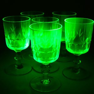 Può includere: Sei calici vintage in vetro verde con motivi floreali incisi. I bicchieri hanno un gambo corto e una base rotonda. I calici sono disposti in un motivo circolare su uno sfondo scuro, illuminati da una luce verde brillante.