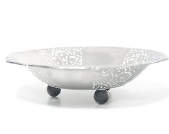 May include: A silver-colored decorative bowl with a patterned rim and three dark gray ball feet. The bowl has a shallow, oval shape and a textured design on the sides.