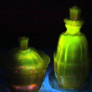 May include: Two antique, glowing green glass containers. One is a lidded jar, the other a bottle with a stopper. The glass has a unique, textured appearance, and the objects are illuminated with a bright, neon-like glow.