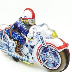 May include: A vintage tin toy motorcycle featuring a police officer. The motorcycle is predominantly white with red and blue detailing. The officer is dressed in a blue uniform and a red and white helmet, marked with "P.D.". The wheels are a dark brown colour.